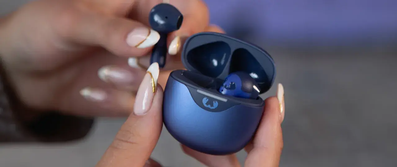 Une main aux ongles soignés tient un écouteur bleu qui semble trop grand pour l'oreille. L'étui de chargement est ouvert, révélant un autre écouteur à l'intérieur.