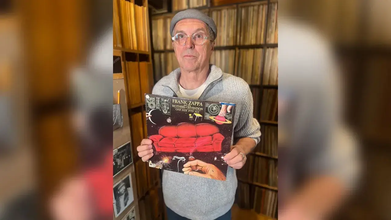 Un homme tient la pochette de l'album "One Size Fits All" de Frank Zappa, entouré de vinyles.
