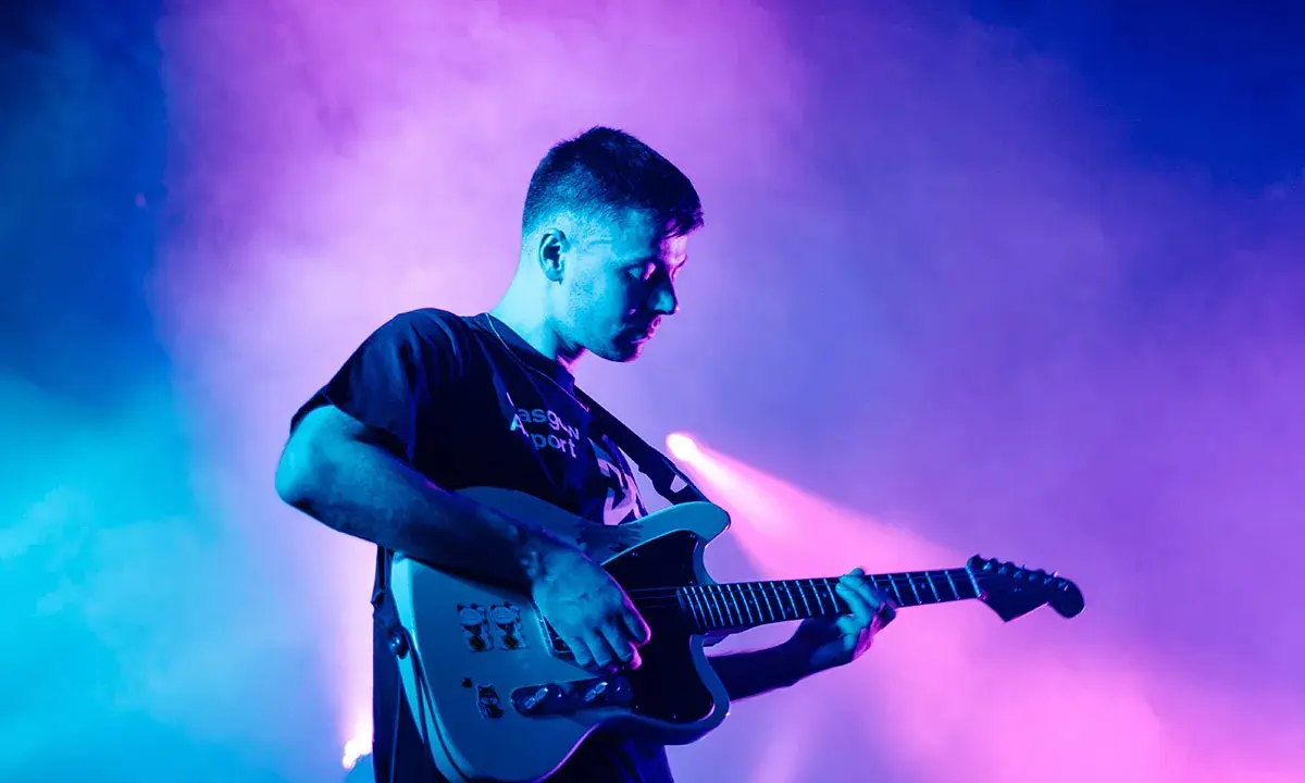 Un jeune homme joue de la guitare électrique sous des lumières bleues et roses. Il est en train de trouver des musiciens pour son groupe.