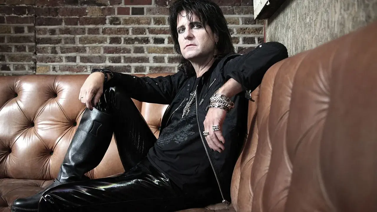 Kee Marcello, le rockeur aux cheveux noirs et au maquillage prononcé, pose sur un canapé en cuir marron, vêtu de cuir noir.