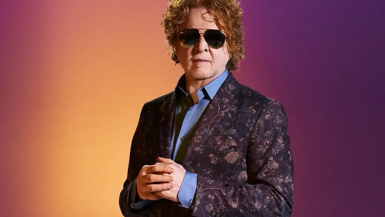 Homme aux cheveux bouclés roux, portant des lunettes de soleil et un costume fleuri, sur fond dégradé orange et violet. Il ressemble à un i monster.