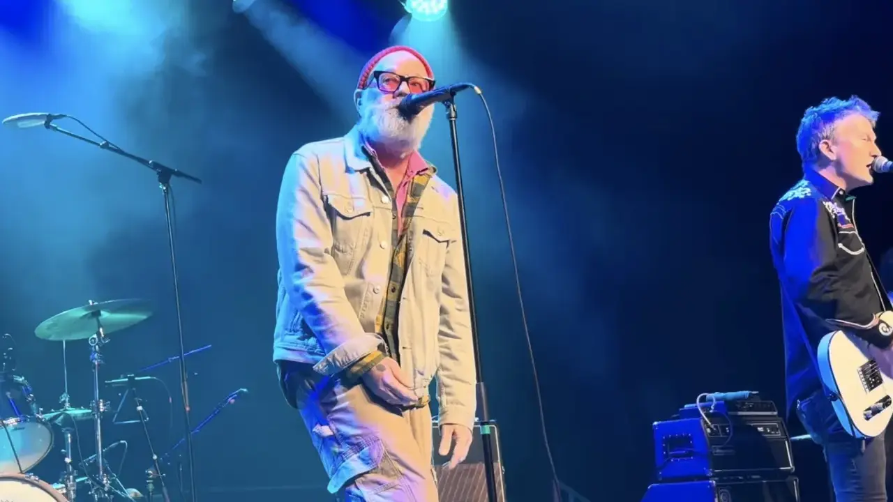 Le chanteur principal du rem groupe, avec une barbe blanche et un bonnet rouge, chante sur scène. Un guitariste est à droite.