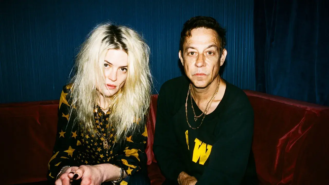 Un couple, aux allures rock, pose sur un canapé rouge. Elle, aux cheveux blonds décoiffés, porte une robe à étoiles. Lui, au regard intense, arbore un sweat noir. Ils semblent vouloir "kill the young" par leur style.