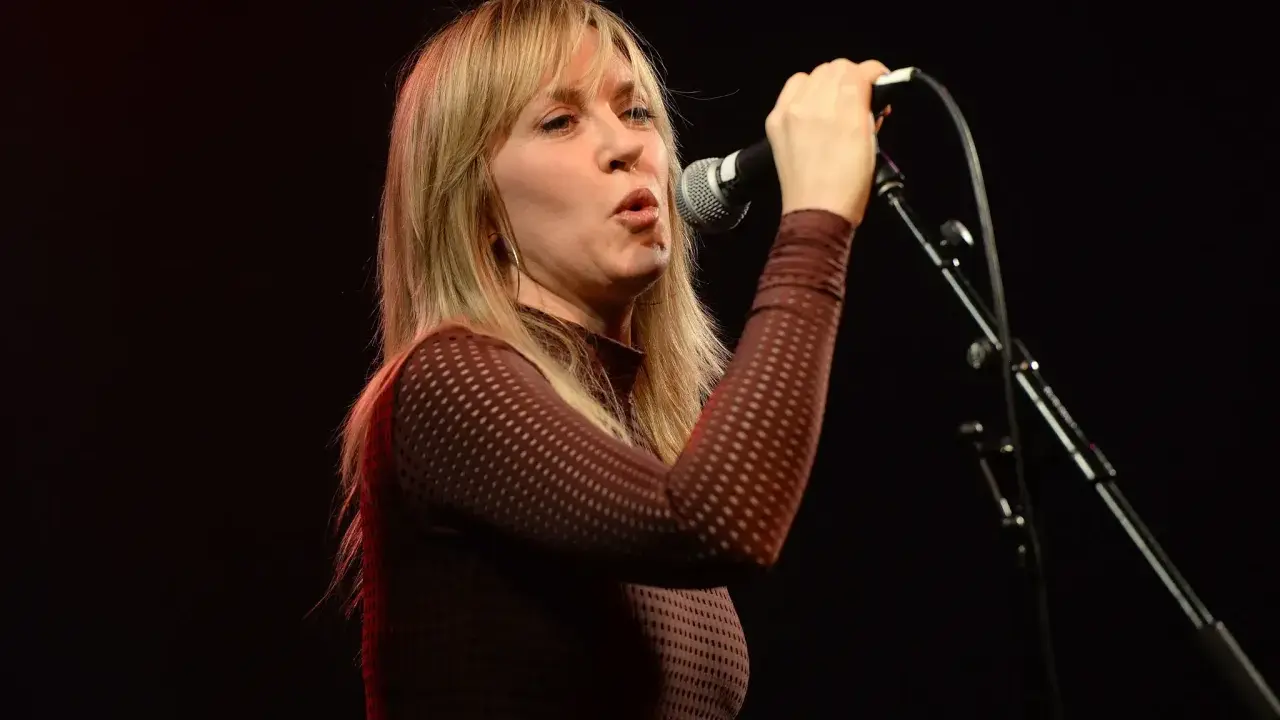 Liz Phair chante avec passion dans un micro, ses cheveux blonds encadrant son visage expressif.