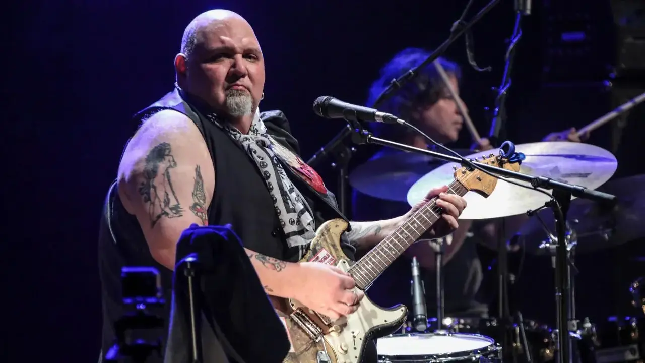Un guitariste popa chubby, chauve, tatoué, joue sur scène avec un batteur en arrière-plan.