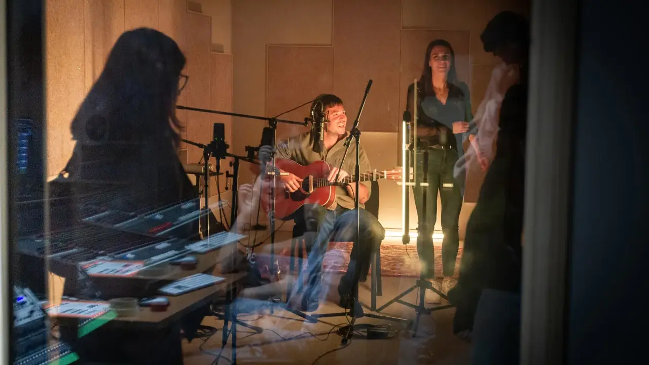 Un homme joue de la guitare dans un studio d'enregistrement, entouré de microphones. Une femme chante à ses côtés. La scène suggère une collaboration musicale, peut-être pour une liste producteur de musique.