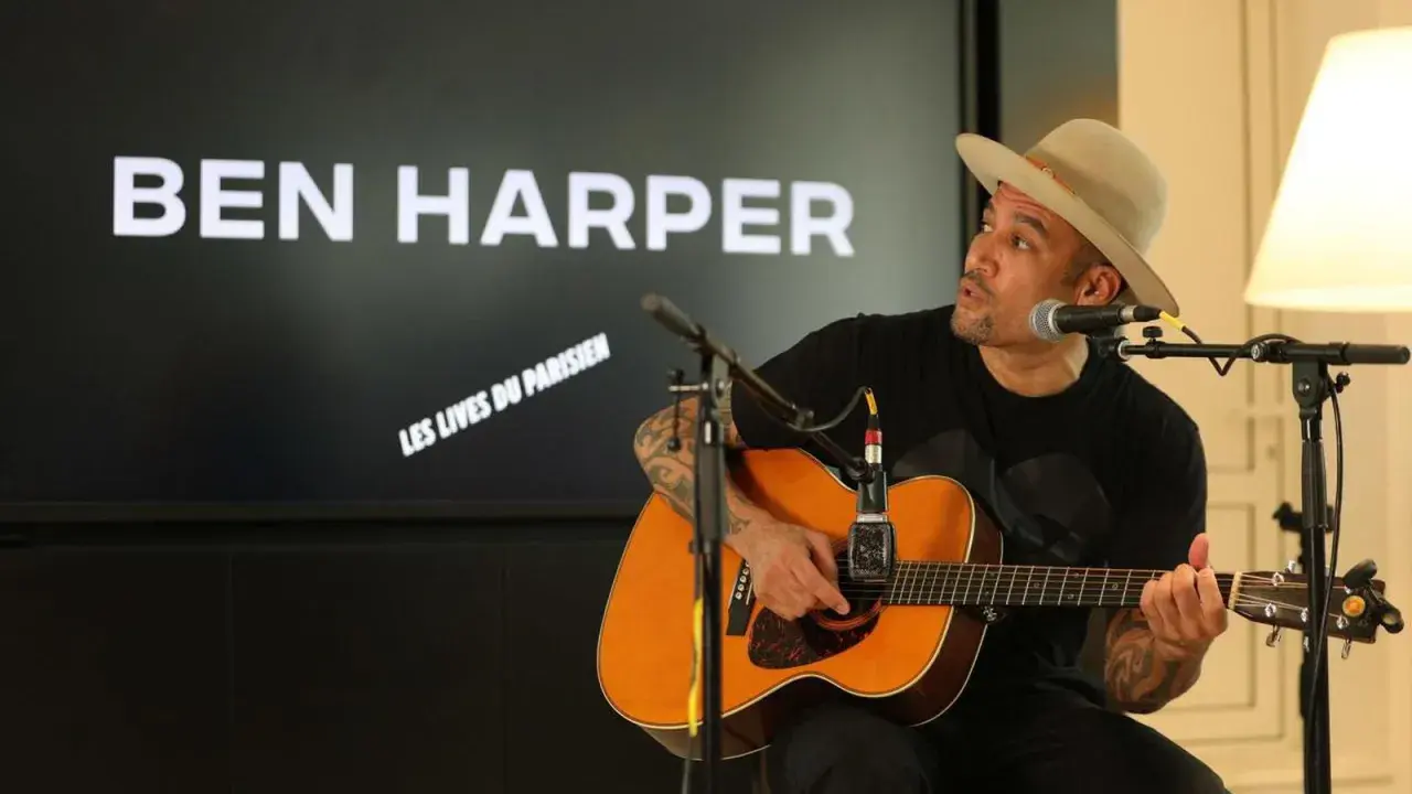 Ben Harper, chapeau sur la tête, joue de la guitare devant un écran affichant son nom et "Les Vies du Parisien".