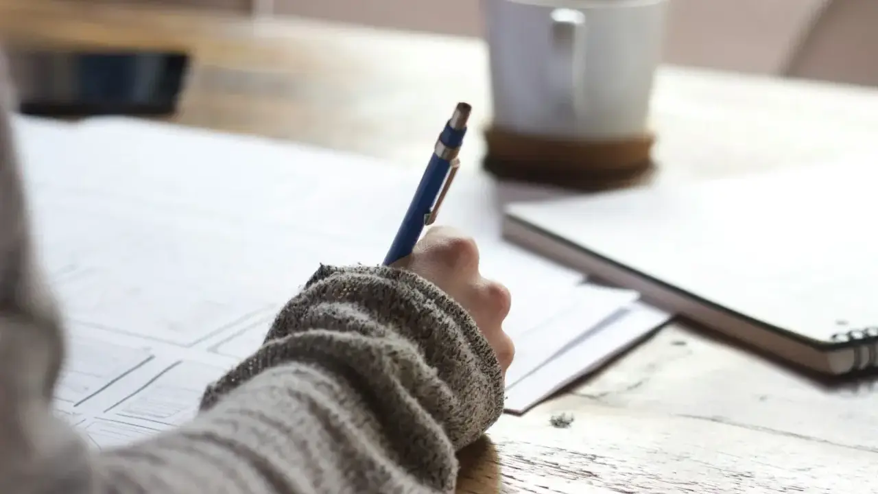 Main tenant une plume bleue, une personne écrit sur du papier, une tasse de café à proximité. Une idée de chanson à écrire prend forme.