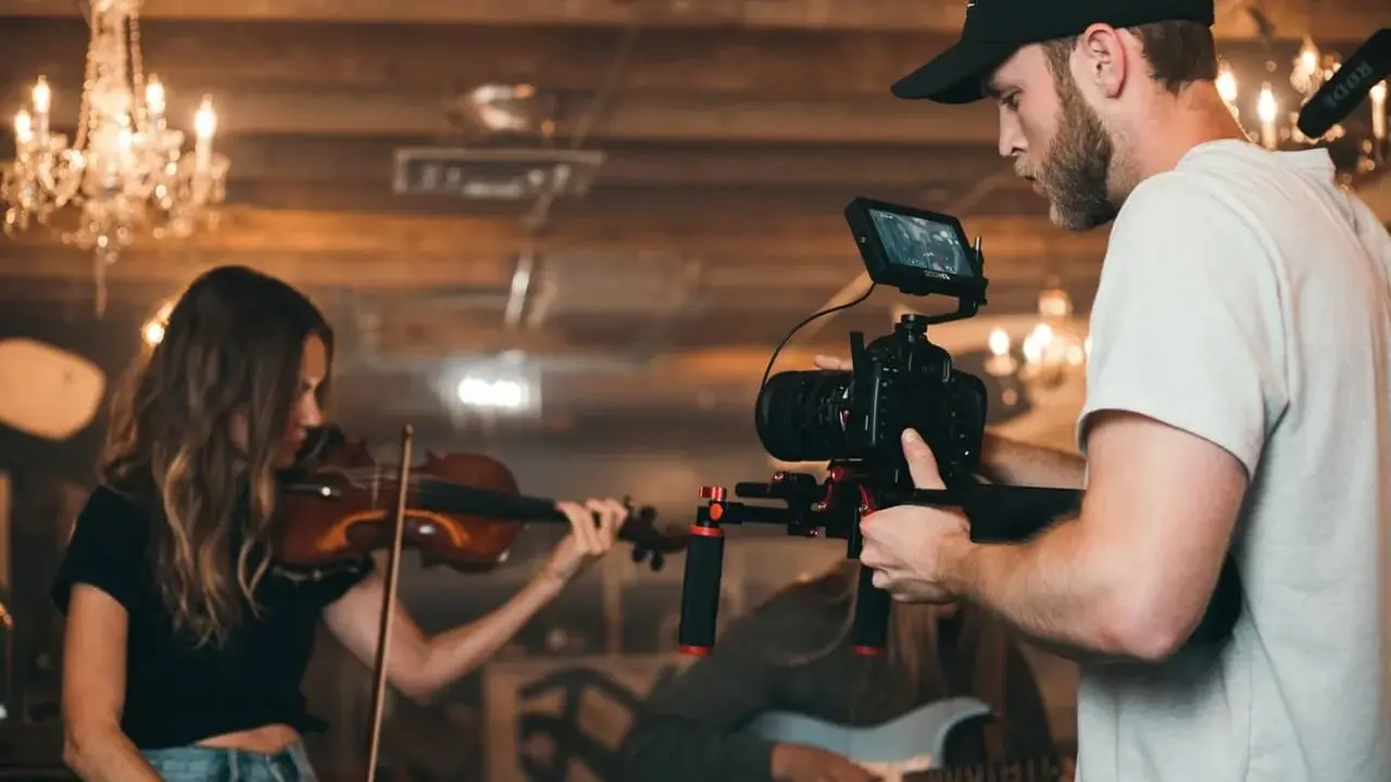 Un caméraman s'apprête à créer un clip vidéo, filmant une violoniste et un guitariste dans un décor chaleureux avec des lustres.