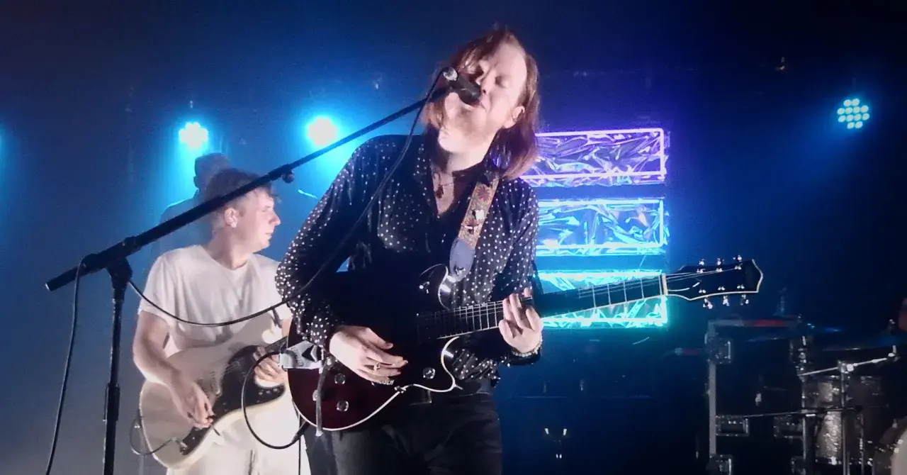 Le groupe Two Doors Cinema Club en concert, éclairé par des néons bleus. Le chanteur joue de la guitare et chante dans un micro.
