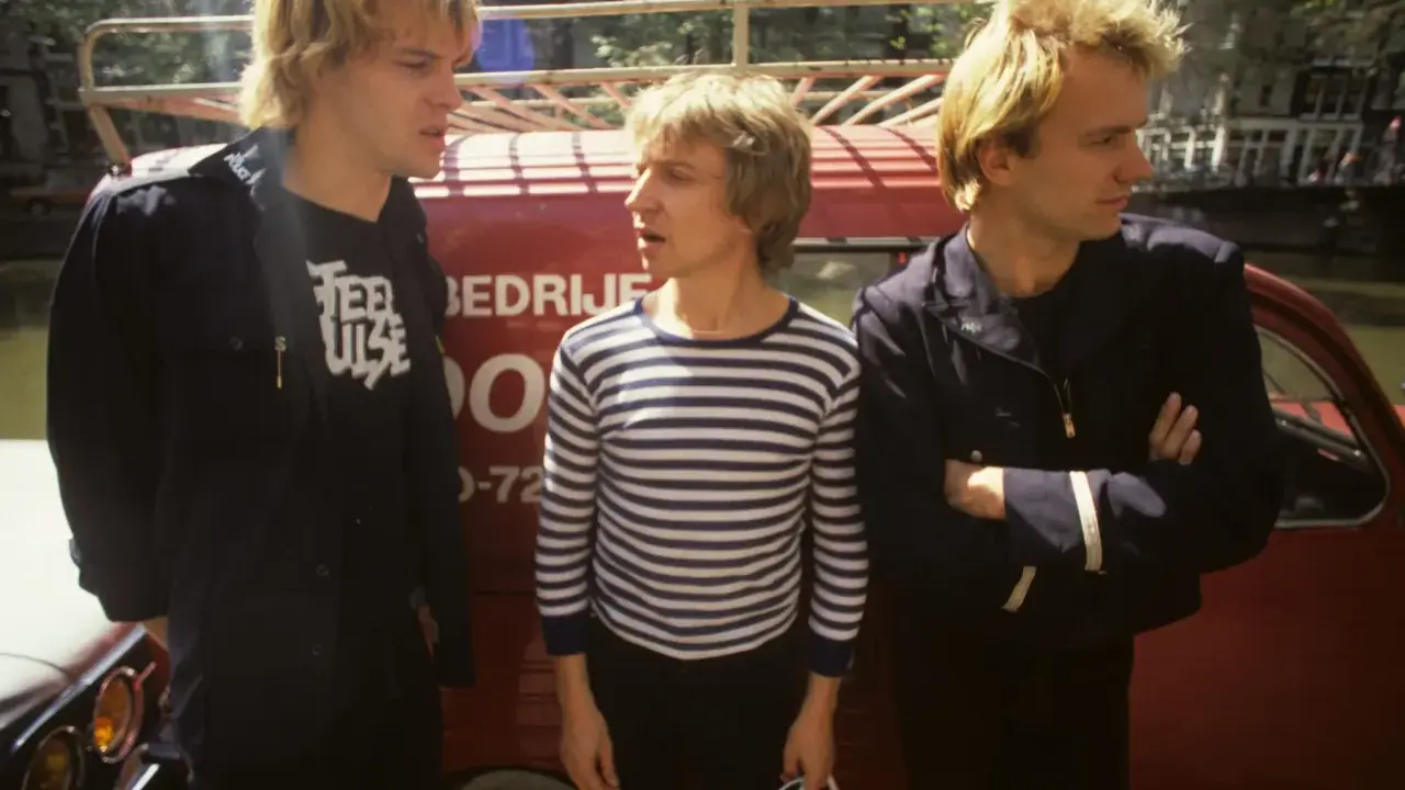 Le groupe Police, dans les années 80, pose devant un véhicule rouge. L'un porte un t-shirt à rayures, les autres des vestes sombres.