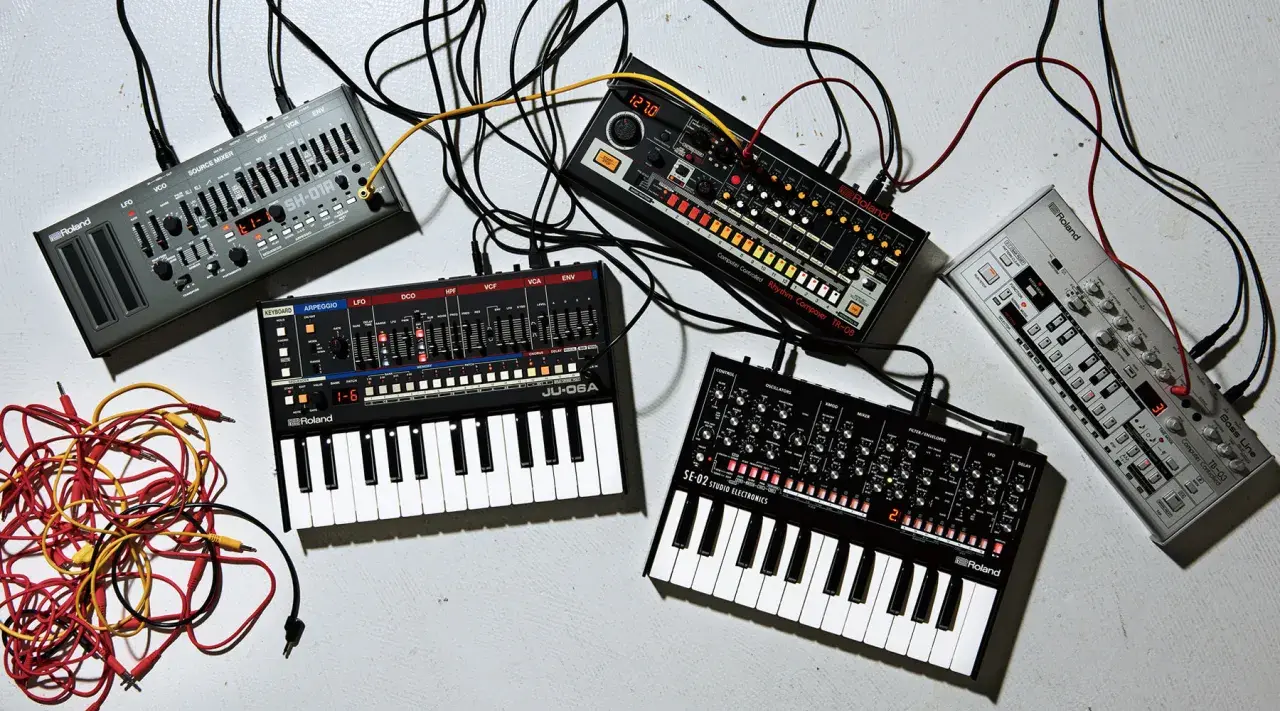 Plusieurs petits instruments de musique électroniques, dont des synthétiseurs et des boîtes à rythmes, sont connectés par des câbles.
