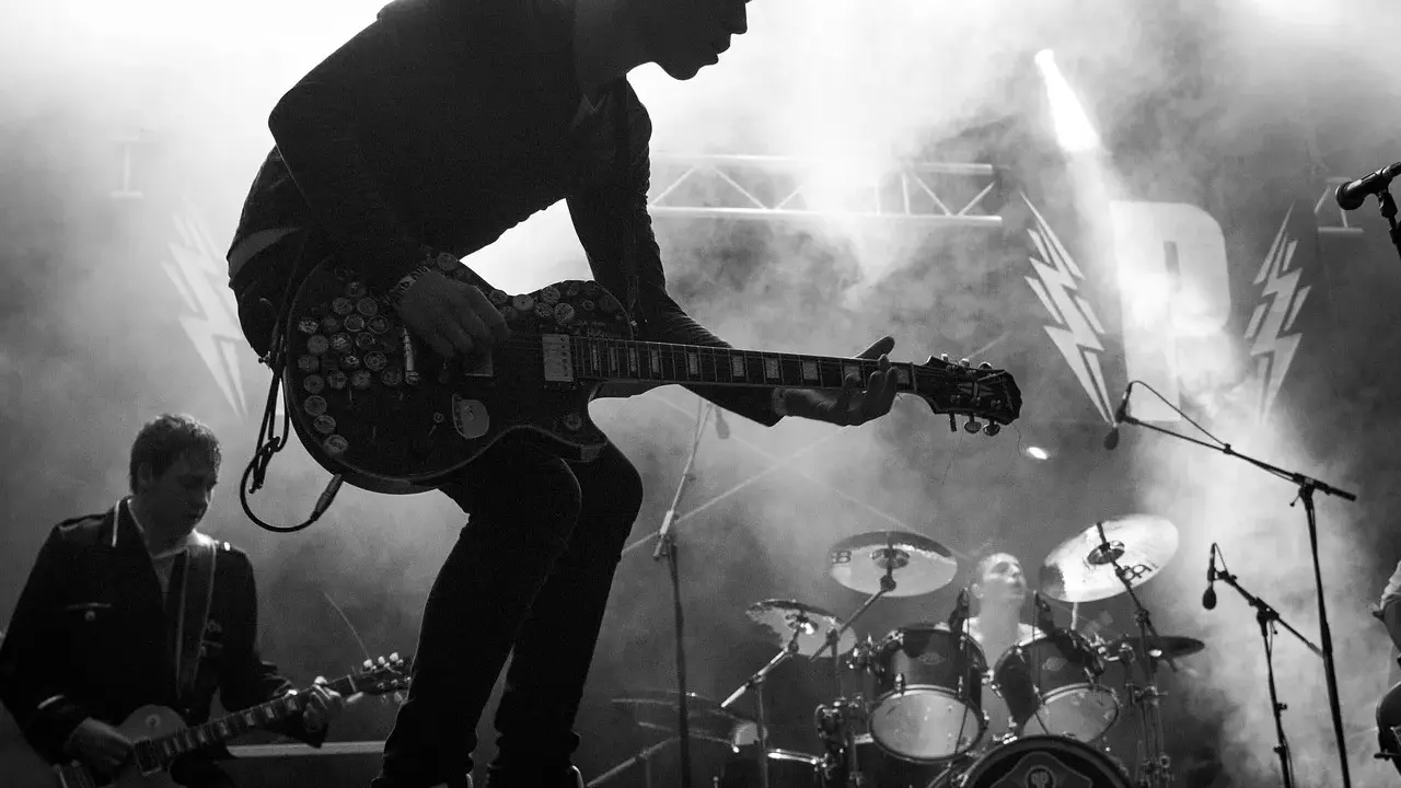 Un guitariste électrique joue sur scène, entouré de fumée. Derrière lui, un batteur et le logo d'un nom de groupe de rock.