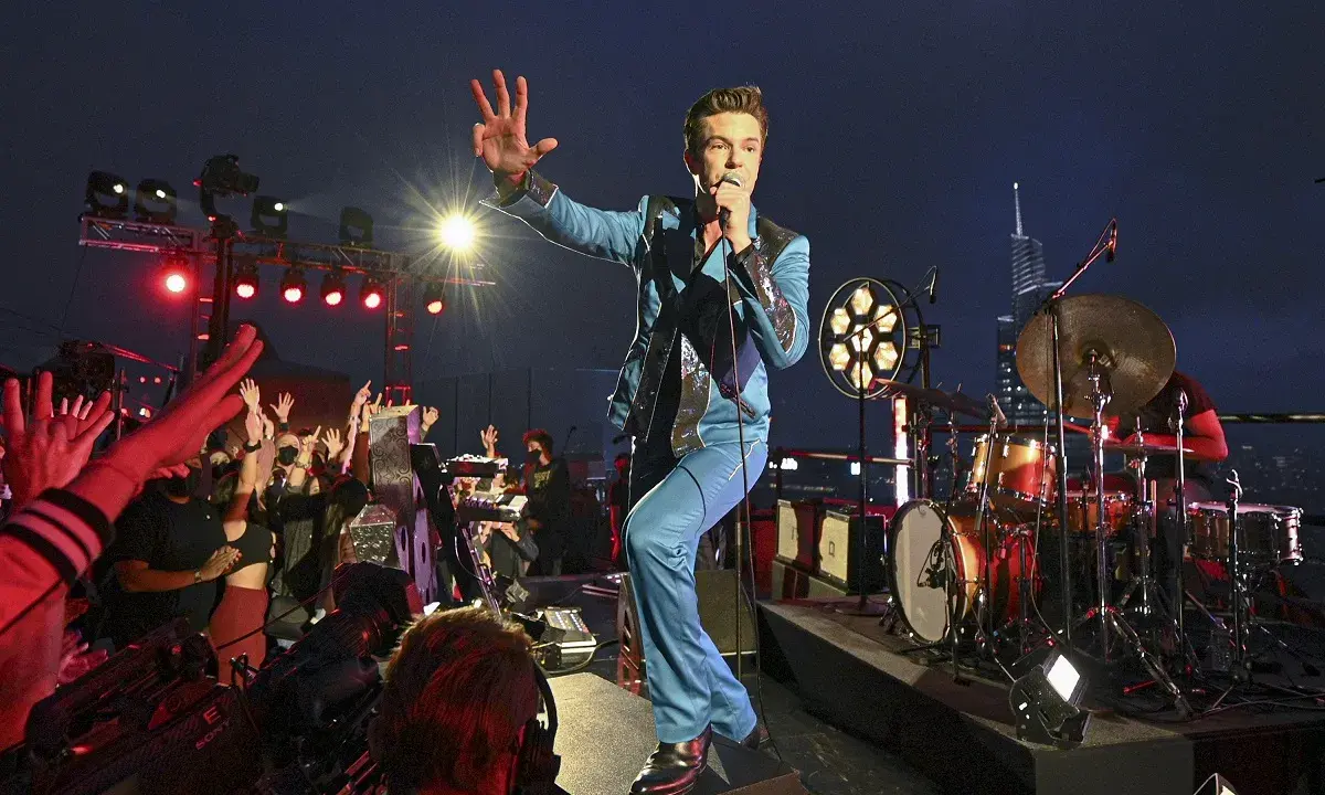Brandon Flowers de The Killers, en costume bleu scintillant, salue la foule lors d'un concert. Les albums de The Killers résonnent dans la nuit.