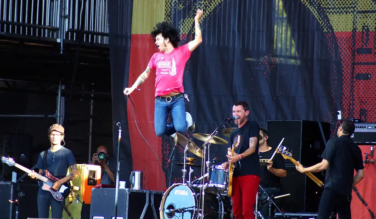 Un chanteur saute sur scène, micro en main, lors d'un concert en plein air. Le groupe joue avec énergie, une ambiance de festival.