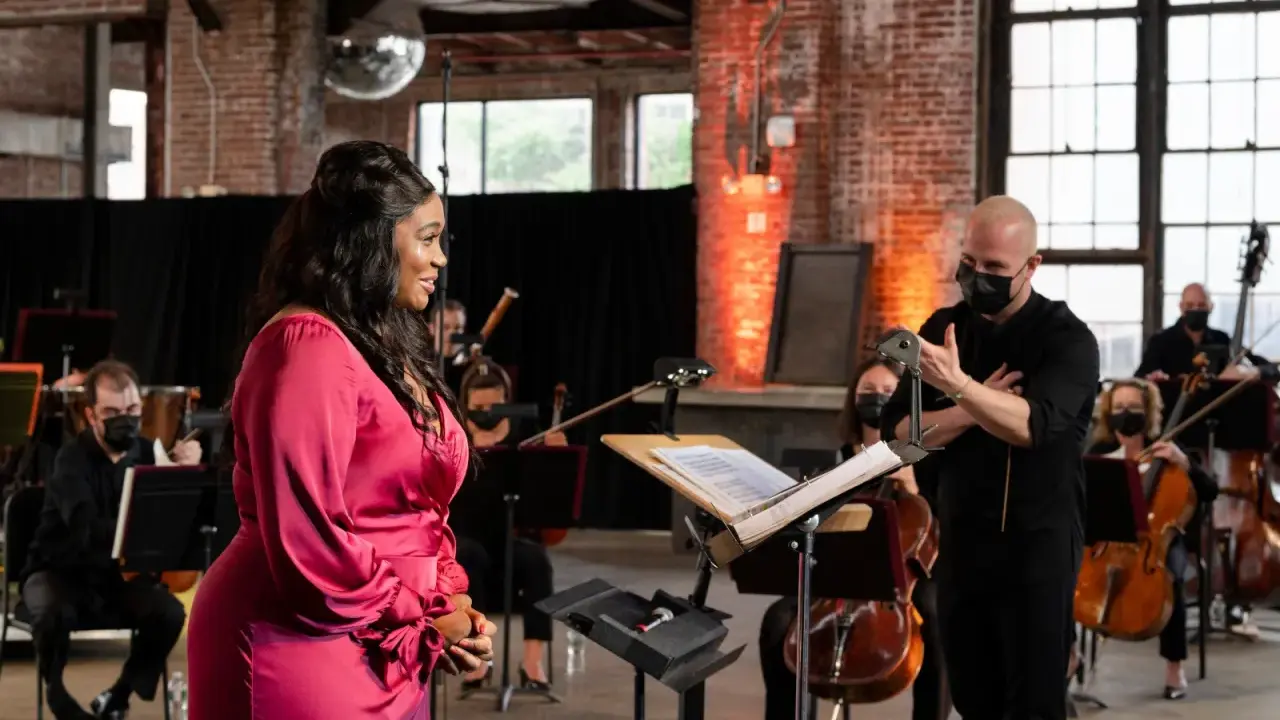 Une chanteuse en robe rose discute avec un chef d'orchestre masqué, entourée de musiciens. Elle cherche peut-être à trouver un agent pour sa carrière.