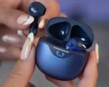 Une main aux ongles soignés tient un écouteur bleu qui semble trop grand pour l'oreille. L'étui de chargement est ouvert, révélant un autre écouteur à l'intérieur.