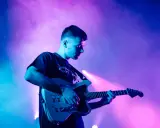 Un jeune homme joue de la guitare électrique sous des lumières bleues et roses. Il est en train de trouver des musiciens pour son groupe.