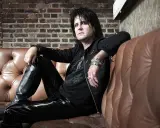Kee Marcello, le rockeur aux cheveux noirs et au maquillage prononcé, pose sur un canapé en cuir marron, vêtu de cuir noir.