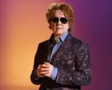 Homme aux cheveux bouclés roux, portant des lunettes de soleil et un costume fleuri, sur fond dégradé orange et violet. Il ressemble à un i monster.