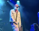 Le chanteur principal du rem groupe, avec une barbe blanche et un bonnet rouge, chante sur scène. Un guitariste est à droite.