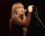 Liz Phair chante avec passion dans un micro, ses cheveux blonds encadrant son visage expressif.