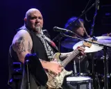 Un guitariste popa chubby, chauve, tatoué, joue sur scène avec un batteur en arrière-plan.