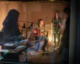 Un homme joue de la guitare dans un studio d'enregistrement, entouré de microphones. Une femme chante à ses côtés. La scène suggère une collaboration musicale, peut-être pour une liste producteur de musique.