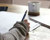 Main tenant une plume bleue, une personne écrit sur du papier, une tasse de café à proximité. Une idée de chanson à écrire prend forme.