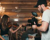 Un caméraman s'apprête à créer un clip vidéo, filmant une violoniste et un guitariste dans un décor chaleureux avec des lustres.