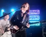 Le groupe Two Doors Cinema Club en concert, éclairé par des néons bleus. Le chanteur joue de la guitare et chante dans un micro.