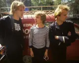 Le groupe Police, dans les années 80, pose devant un véhicule rouge. L'un porte un t-shirt à rayures, les autres des vestes sombres.