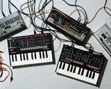 Plusieurs petits instruments de musique électroniques, dont des synthétiseurs et des boîtes à rythmes, sont connectés par des câbles.