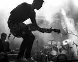 Un guitariste électrique joue sur scène, entouré de fumée. Derrière lui, un batteur et le logo d'un nom de groupe de rock.