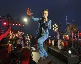 Brandon Flowers de The Killers, en costume bleu scintillant, salue la foule lors d'un concert. Les albums de The Killers résonnent dans la nuit.