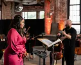 Une chanteuse en robe rose discute avec un chef d'orchestre masqué, entourée de musiciens. Elle cherche peut-être à trouver un agent pour sa carrière.