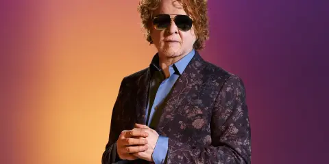 Homme aux cheveux bouclés roux, portant des lunettes de soleil et un costume fleuri, sur fond dégradé orange et violet. Il ressemble à un i monster.