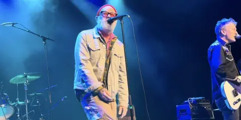 Le chanteur principal du rem groupe, avec une barbe blanche et un bonnet rouge, chante sur scène. Un guitariste est à droite.