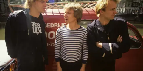 Le groupe Police, dans les années 80, pose devant un véhicule rouge. L'un porte un t-shirt à rayures, les autres des vestes sombres.