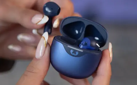 Une main aux ongles soignés tient un écouteur bleu qui semble trop grand pour l'oreille. L'étui de chargement est ouvert, révélant un autre écouteur à l'intérieur.