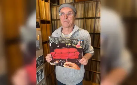 Un homme tient la pochette de l'album "One Size Fits All" de Frank Zappa, entouré de vinyles.