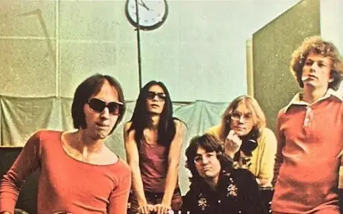 Cinq membres des Flamin' Groovies posent dans un studio. L'un d'eux porte des lunettes de soleil et un t-shirt orange.