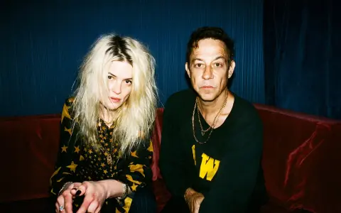 Un couple, aux allures rock, pose sur un canapé rouge. Elle, aux cheveux blonds décoiffés, porte une robe à étoiles. Lui, au regard intense, arbore un sweat noir. Ils semblent vouloir "kill the young" par leur style.