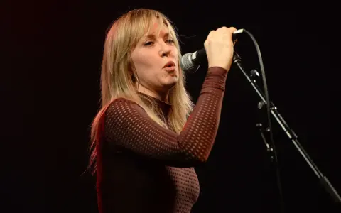Liz Phair chante avec passion dans un micro, ses cheveux blonds encadrant son visage expressif.