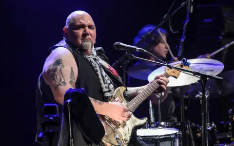 Un guitariste popa chubby, chauve, tatoué, joue sur scène avec un batteur en arrière-plan.