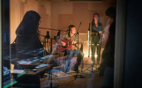 Un homme joue de la guitare dans un studio d'enregistrement, entouré de microphones. Une femme chante à ses côtés. La scène suggère une collaboration musicale, peut-être pour une liste producteur de musique.