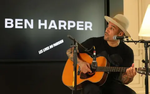 Ben Harper, chapeau sur la tête, joue de la guitare devant un écran affichant son nom et "Les Vies du Parisien".
