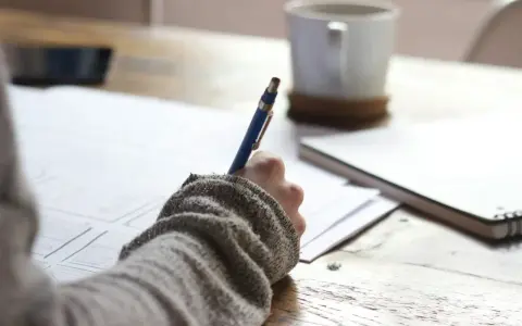 Main tenant une plume bleue, une personne écrit sur du papier, une tasse de café à proximité. Une idée de chanson à écrire prend forme.