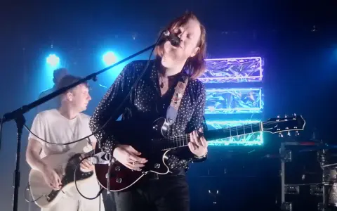 Le groupe Two Doors Cinema Club en concert, éclairé par des néons bleus. Le chanteur joue de la guitare et chante dans un micro.