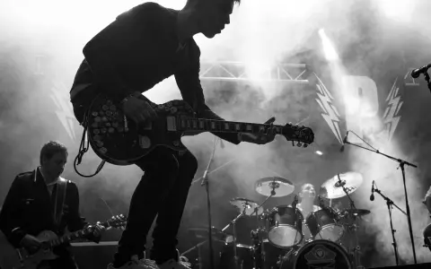 Un guitariste électrique joue sur scène, entouré de fumée. Derrière lui, un batteur et le logo d'un nom de groupe de rock.